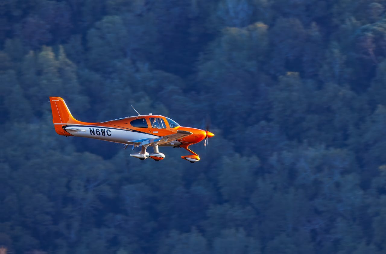 about-02 Bright orange aircraft soaring over a lush green forest on a clear day.