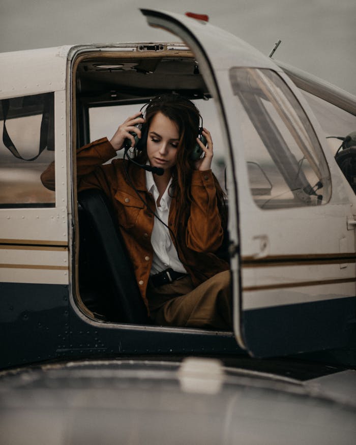 our-experience Charming female with long hair in trendy wear and headset sitting in cockpit of modern airplane with opened door in airfield